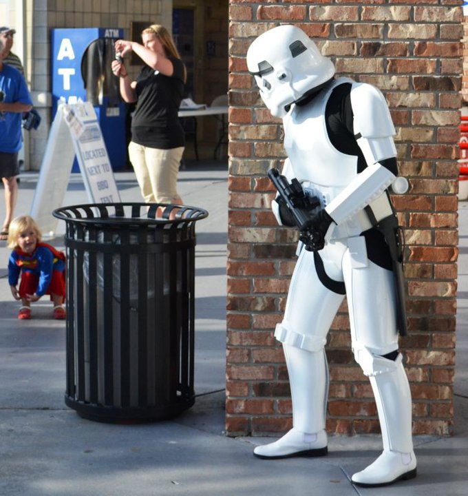 A little girl dressed as Supergirl is looking up at a Stormtrooper cosplayer pretending to hide behind a wall